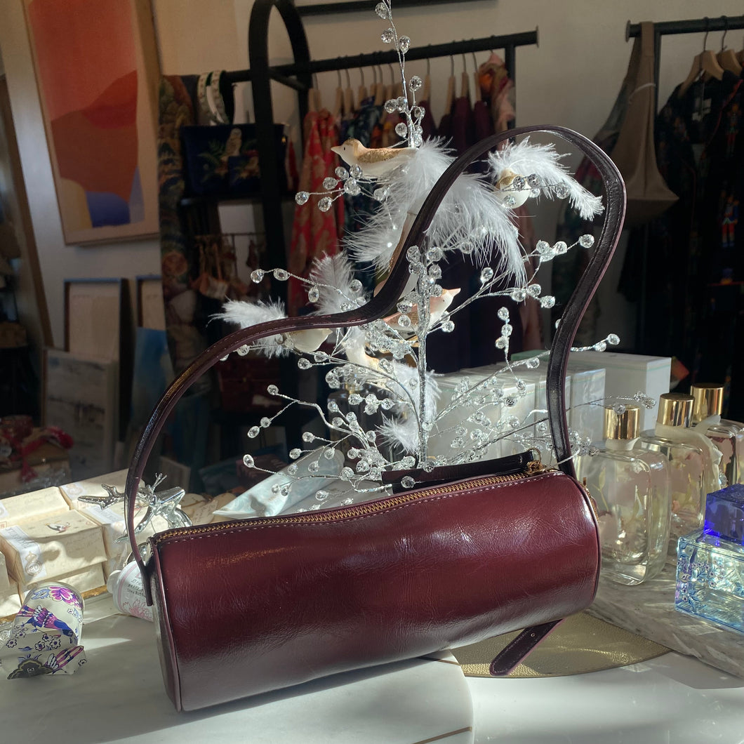 Dark brown vegan leather cylindrical shoulder bag displayed in a boutique setting, resting among holiday decor with white feathers, crystal sprigs, and perfume bottles in the background.