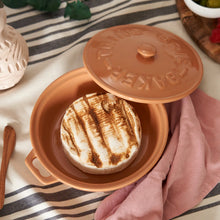 Load image into Gallery viewer, Terracotta-colored stoneware brie baker with handles holding baked brie, shown with the matching lid, wooden spreader, and pink napkin resting on a striped cloth.   