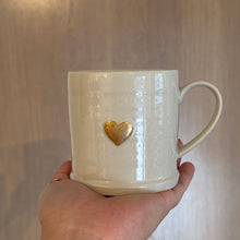 Load image into Gallery viewer, Hand holding a white stoneware mug with a gold heart design against a wood background.