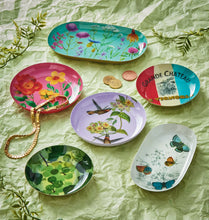 Load image into Gallery viewer, Assorted vintage-style metal trinket dishes in various shapes and designs including round purple hummingbird, round pink floral, oblong white butterfly, oblong teal floral, small round green grapes, and small round red, white, and blue Grande Chateau dish, displayed on a light green textured surface with gold coins, a gold necklace, and greenery accents. 