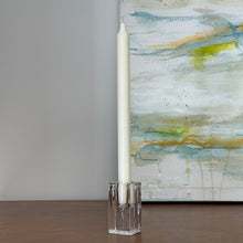 Load image into Gallery viewer, Chunky rectangular clear pressed glass taper candle holder with white taper candle, displayed on wood surface with abstract artwork. 