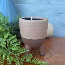 Load image into Gallery viewer, Footed Citro Candle Pot rustic terracotta with terracotta-colored upper half and brown bottom, hand poured citronella candle with single wick in three leg footed pot on wooden surface with greenery. 