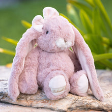 Load image into Gallery viewer, Friendly Kanini soft pink bunny plushie with long floppy ears , white muzzle, small  pink nose, pale pink bow on ear and paw pads seated on a stone surface with green foliage in the background. 