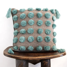 Load image into Gallery viewer, Jitana handcrafted wool throw pillow in gray with blue pom-pom accents and corner tassels, displayed on a wooden stool on a white background. 