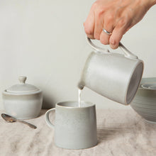 Load image into Gallery viewer, Aquarius Sage stoneware creamer with reactive glaze, pouring into matching mug on a cloth surface with coordinating sugar bowl and bowls.