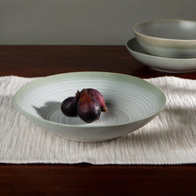 Load image into Gallery viewer, Aquarius Sage stoneware pasta serving bowl with reactive glaze and concentric line pattern, on a wood surface with figs and a white placemat. 