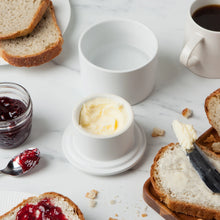 Load image into Gallery viewer, Matte round white stoneware butter crock with butter and lid on a marble surface with bread, jam, and coffee. 