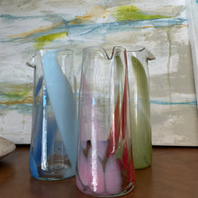 Load image into Gallery viewer, Three tall clear glass pitchers with elongated color accents-blue and light blue, light pink and red, and pistachio green- each with a pour spout, displayed on a wooden surface in front of abstract painting. 