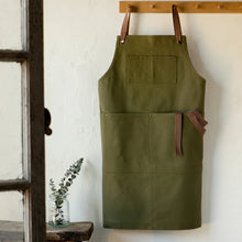 Load image into Gallery viewer, Olive Branch Mason cotton apron in olive green with front pockets, brown straps, and metal grommets, displayed hanging from wooden coat rack on a white wall. 