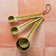 Load image into Gallery viewer, Brio enameled steel measuring spoons with green and gold block print leaf pattern and matte gold finish, set of four, linked with gold chain, displayed on a pink tile surface.
