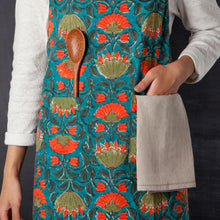 Load image into Gallery viewer, Allure Block Print Apron, cotton apron with hand block-printed floral pattern in orange and olive on a teal background, featuring  side-by-side front pockets with a wooden spoon and towel, displayed on a model against a dark background.