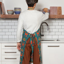 Load image into Gallery viewer, Allure Block Print Apron, cotton apron with hand block-printed floral pattern in coral and olive on a teal background, featuring waist ties, shown from the back on a model in a kitchen setting. 