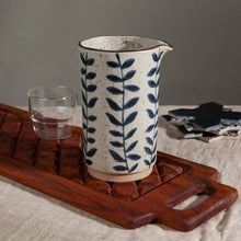 Load image into Gallery viewer, Vine Element Pitcher, stoneware pitcher with reactive glaze and blue vine pattern on cream speckled surface, beige base, and brown rim with spout, displayed on a wooden board with glass in the background. 