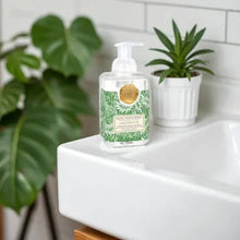 Load image into Gallery viewer, Michel Design Works Greenhouse foaming shea butter hand soap pump bottle with gold seal and green botanical design displayed on a white sink with potted plants in the background. 