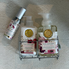 Load image into Gallery viewer, Michel Design Works Pressed Flowers shea butter foaming hand wash and body lotion in clear pump bottles with pink, purple, and cream floral labels and gold seal accents, displayed in a double-compartment metal caddy with matching room spray in metal silver bottle, on a neutral textured fabric surface. 