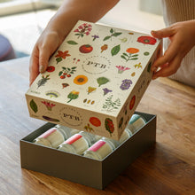 Load image into Gallery viewer, Plant the Box Votive Gift Set, KOBO set with illustrated botanical lid featuring multicolored flowers, leaves, and fruit, box being opened to reveal multiple glass jar votive candles inside a dark box base, held by hands over a wooden surface with soft indoor background. 
