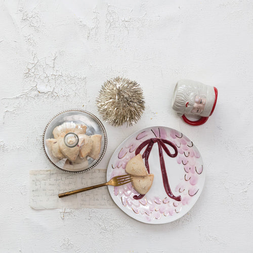 Round white ceramic dish with a pink and red wreath and bow design. Gold fork and two cookies on dish; next to a glass ,silver rimmed cloche with cookies inside; a gold pom pom; and a red and white Santa mug. On a white textured surface. 