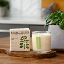 Load image into Gallery viewer, Fresh Picked Basil Plant The Box Candle, unlit white soy wax candle in clear glass jar with green label beside illustrated basil plant product box on wooden board with potted basil plant in background. 