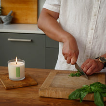Load image into Gallery viewer, Fresh Picked Basil Plant The Box Candle, lit white soy wax candle in clear glass jar with green label beside person chopping fresh basil on wooden board in kitchen setting. 