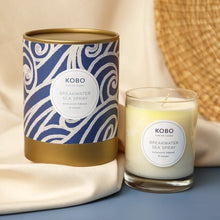 Load image into Gallery viewer, Breakwater Sea Spray Coastal Candle, KOBO pure soy candle in clear glass jar with cream wax beside blue patterned packaging with white wave-inspired swirl pattern and gold-toned rim and base, displayed on blue surface with beige fabric against soft neutral background. 