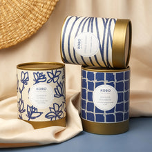 Load image into Gallery viewer, Citrus Sunset Coastal Candle, cream packaging with blue horizontal stripe pattern and gold-toned rim and base stacked on two coordinating patterned packages, displayed on blue surface with draped beige fabric against soft neutral background. 