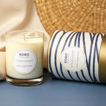 Load image into Gallery viewer, Citrus Sunset Coastal Candle, KOBO pure soy candle in clear glass jar with cream wax beside cream packaging with blue horizontal stripe pattern and gold-toned rim and base lying on its side, displayed on blue surface against soft neutral background. 