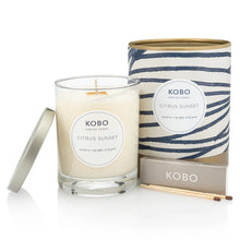 Load image into Gallery viewer, Citrus Sunset Coastal Candle, KOBO pure soy candle in clear glass jar with cream wax and silver-toned lid resting at the side, cream packaging with blue horizontal stripe pattern and gold-toned rim and base beside, KOBO gray matchbox and two brown-tipped wooden matches in front, on white background. 