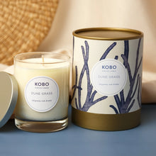 Load image into Gallery viewer, Dune Grass Coastal Candle, KOBO pure soy candle in clear glass jar with cream wax and silver-toned lid resting at the side, cream packaging with blue branch pattern and gold-toned rim and base beside, displayed on a blue surface with soft neutral background. 