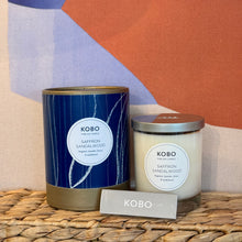 Load image into Gallery viewer, Saffron Sandalwood Coastal Candle, KOBO pure soy candle in clear glass jar with cream wax and silver-toned lid beside blue patterned packaging with gold-toned rim and base, KOBO gray matchbox in front, displayed on woven surface against abstract multicolor background. 