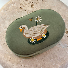 Load image into Gallery viewer, Garden Goose Oval Jewelry Box, olive green oval case with hand-embroidered goose, daisy flowers, and bee motif with subtle bead embellishment, zip closure, on textured stone surface. 