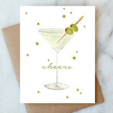Load image into Gallery viewer, Martini Cheers Card, hand-painted watercolor martini glass with green olive garnish and gold foil stars, gold foil script text, on felt textured white card, recycled brown kraft envelope behind, on white marble surface.  
