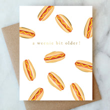 Load image into Gallery viewer, A Weenie Bit Older Card, hand-painted watercolor hot dog pattern with mustard and gold foil accents on textured white card, recycled brown kraft envelope behind, on white marble surface. 