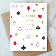 Load image into Gallery viewer, Wishing You A Lucky Year Card, hand-painted white card with gold foil lettering and scattered playing card suit icons in red, black, and gold, recycled brown kraft envelope behind, on white marble surface. 