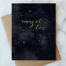 Load image into Gallery viewer, Mazel Tov Celebration Card, hand-painted watercolor dark sky with gold foil script and scattered stars, recycled brown kraft envelope behind, on white marble surface. 