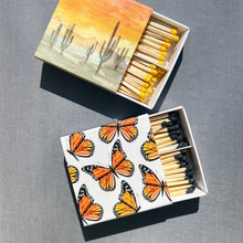 Load image into Gallery viewer, Monarch Boxed Matches, two boxed wooden safety matches featuring watercolor designs, one with orange monarch butterflies on white sliding cover with black tipped-sticks and the other with a desert cactus design with yellow-tipped sticks, partially open on gray surface. 