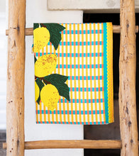 Load image into Gallery viewer, Kitchen Tea Towel, white kitchen tea towel with yellow lemon illustrations, dark green leaves, yellow, teal, coral, and white plaid background, teal and yellow border accents, and yellow rickrack trim, displayed hanging on a rustic wooden ladder against a white background. 