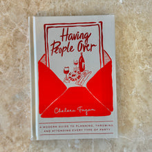 Load image into Gallery viewer, Having People Over by Chelsea Fagan, white hardcover entertaining guide with red envelope illustration and table setting sketch on cover, displayed on brown marble surface. 