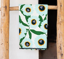 Load image into Gallery viewer, Kitchen Tea Towel, white kitchen towel with oversized light aqua floral prints, green leaves, black flower centers with mustard yellow accents, and navy rickrack trim, displayed hanging on a rustic wooden ladder against a white background. 