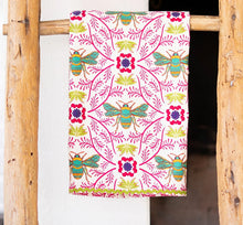Load image into Gallery viewer, Kitchen Tea Towel, white kitchen tea towel with turquoise and yellow bee illustrations, magenta and purple floral motifs, pink botanical vine accents, and light green rickrack trim, displayed hanging on a rustic wooden rack against a white background. 