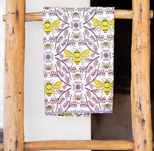 Load image into Gallery viewer, Kitchen Tea Towel, white kitchen tea towel with yellow bee illustrations, lavender  floral motifs, brown botanical vine accents, mint green and reddish orange botanical details, and lilac rickrack trim, displayed hanging on a rustic wooden ladder against a white background. 