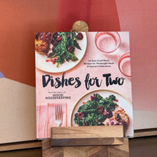 Load image into Gallery viewer, Dishes for Two by Good Housekeeping, soft blush pink illustrated hardcover featuring two plated meals with chicken, greens, and cherries, displayed on wooden stand against geometric framed art background. 