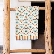 Load image into Gallery viewer, Kitchen Tea Towel, kitchen tea towel with blue eye illustrations, orange and burgundy botanical motifs, mint green background, orange, mint green, and white striped border accents, and blue rickrack trim, displayed hanging on a rustic wooden ladder against a white background. 
