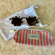 Load image into Gallery viewer, Georgie Cosmic Cowboy Sunglasses, round sunglasses with black, cream, and brown marbled frames and dark brown tinted lenses displayed beside a red-and-light blue vertically striped zip pouch with white "SARDINE" fabric label with yellow stitching, gold fish charm and white microfiber cleaning drawstring storage pouch on brown marble surface.