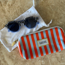 Load image into Gallery viewer, Pool Boy Blues Sunglasses, oversized hexagon sunglasses with black-and-white striped top frames and bridge, light blue lower frames and arms, and dark tinted lenses with 100% UV protection displayed beside red-and-light blue vertically striped zip pouch with white "SARDINE" fabric label with yellow stitching, gold-tone fish charm and white microfiber drawstring cleaning storage bag on brown marble surface. 