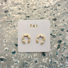 Load image into Gallery viewer, Baguette Horseshoe Studs, pair of handcrafted gold-tone horseshoe-shaped stud earrings with baguette-cut and round clear stones displayed on a white TAI jewelry card on a textured blue-gray surface. 