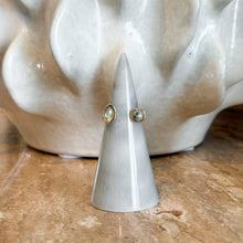 Load image into Gallery viewer, Gold Labradorite Open Ring, handcrafted open- design gold-tone ring with marquise and round gray bezel-set labradorite stones, displayed on a white cone ring holder against a sculptural cream background. 