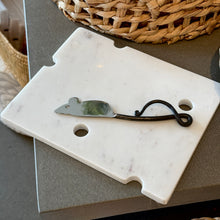 Load image into Gallery viewer, Swiss Cheese Marble Board, hand-polished white marble cheese board with Swiss cheese-inspired cutout details, displayed with a silver mouse-shaped cheese knife on a dark surface beside a woven basket. 