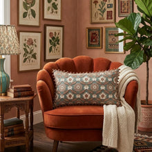 Load image into Gallery viewer, Embroidered Lumbar Pillow, handcrafted teal and mauve pink lumbar pillow with intricate floral embroidery and reddish-brown pom pom trim, displayed on a rust velvet accent chair with a cream knit throw blanket and a decorated gallery wall in the background. 