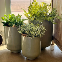 Load image into Gallery viewer, Sage Green Metal Footed Nesting Pots Set, trio of sage green enamel-finish metal nesting pots with warm metallic top rims, displayed with assorted greenery on a concrete floor near a window.