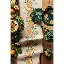 Load image into Gallery viewer, Cedar &amp; Citrus, two dark forest green cloth napkins with a light green leafy and evergreen print tied with a dried orange slice and styled alongside a sliced orange and leaf design table runner and coordinating tableware on a wooden tabletop. 